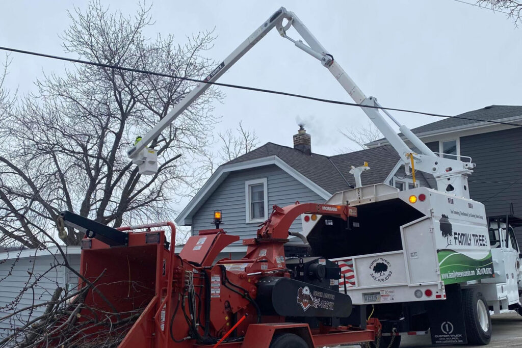 A bucket truck parked in front of a blue house, with a hopper attached containing sticks and debris.